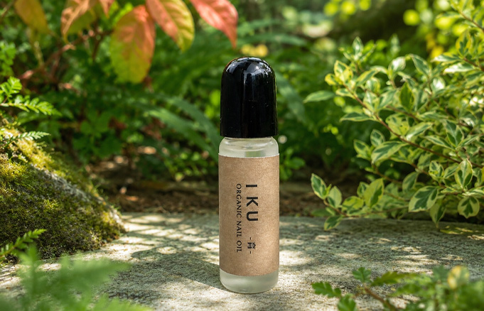 Bottle of IKU Organic Nail Oil displayed outdoors on a stone surface among green plants and mossy ground.