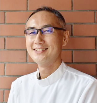 Smiling man with blue glasses and short gray hair in a white coat, posed against a brick wall.
